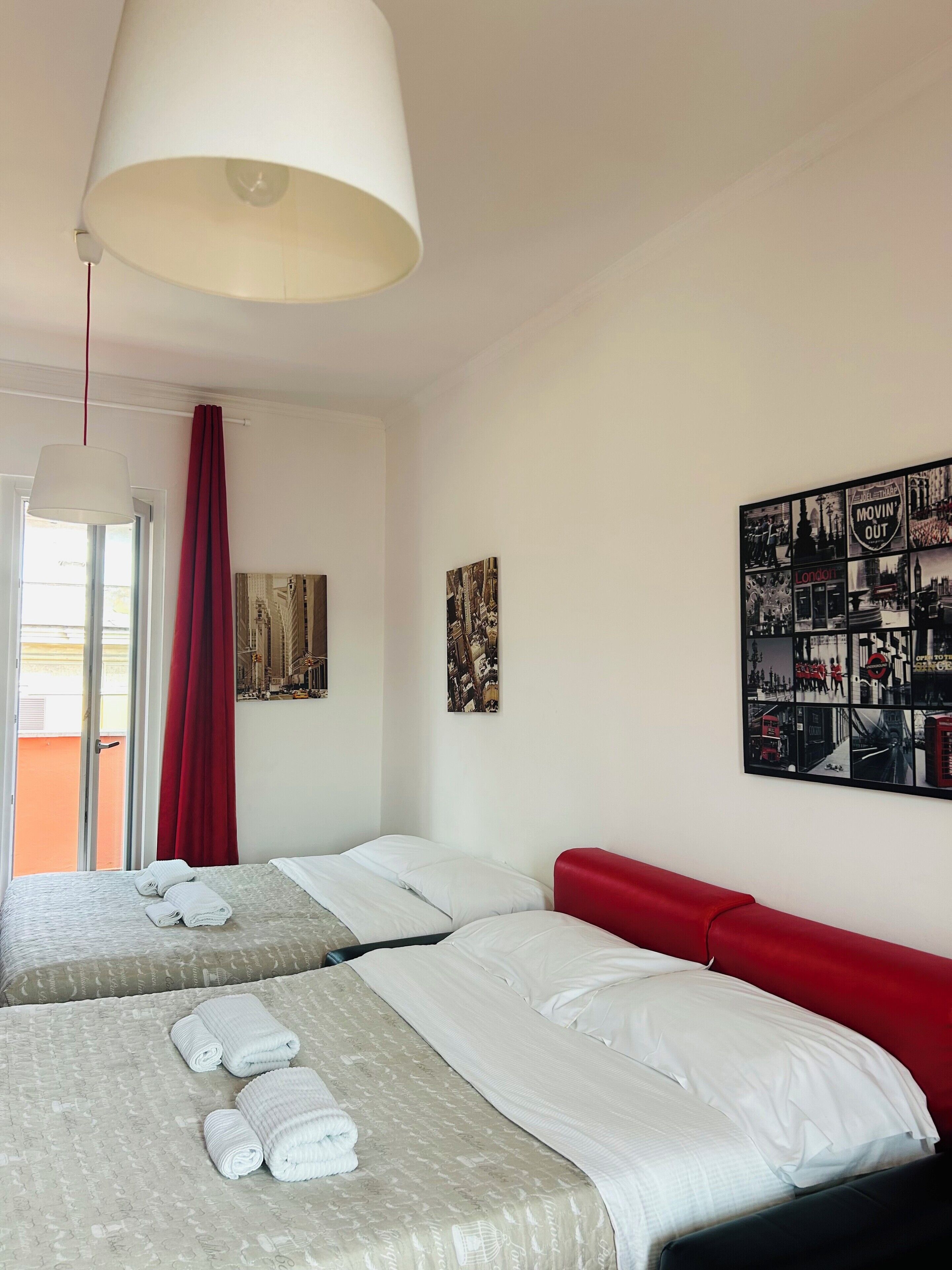 A hotel room with two beds, a red headboard, a large window with red curtains, and a wall with a collage of black and white photos.