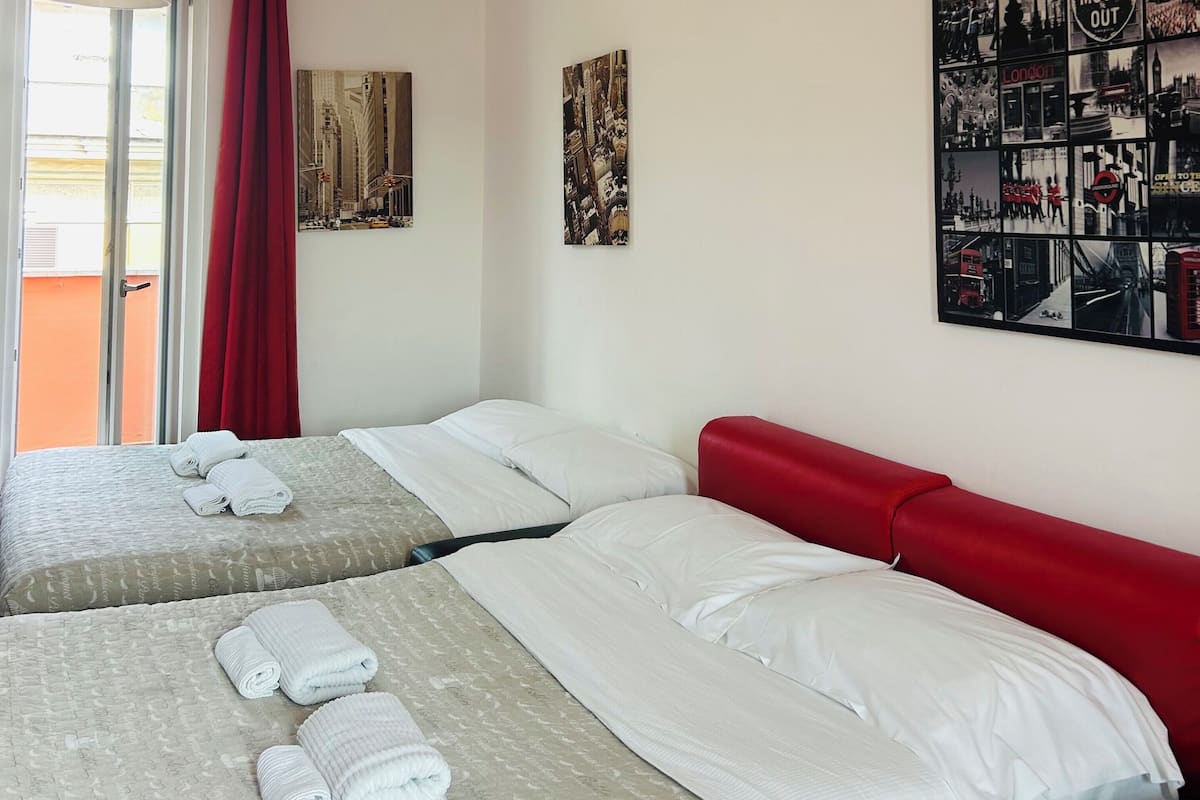 A hotel room with two beds, a red headboard, a large window with red curtains, and a wall with a collage of black and white photos.