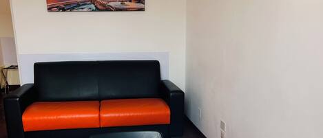 A dining area with a white table and chairs, a black and orange sofa, and a painting of a cityscape on the wall.
