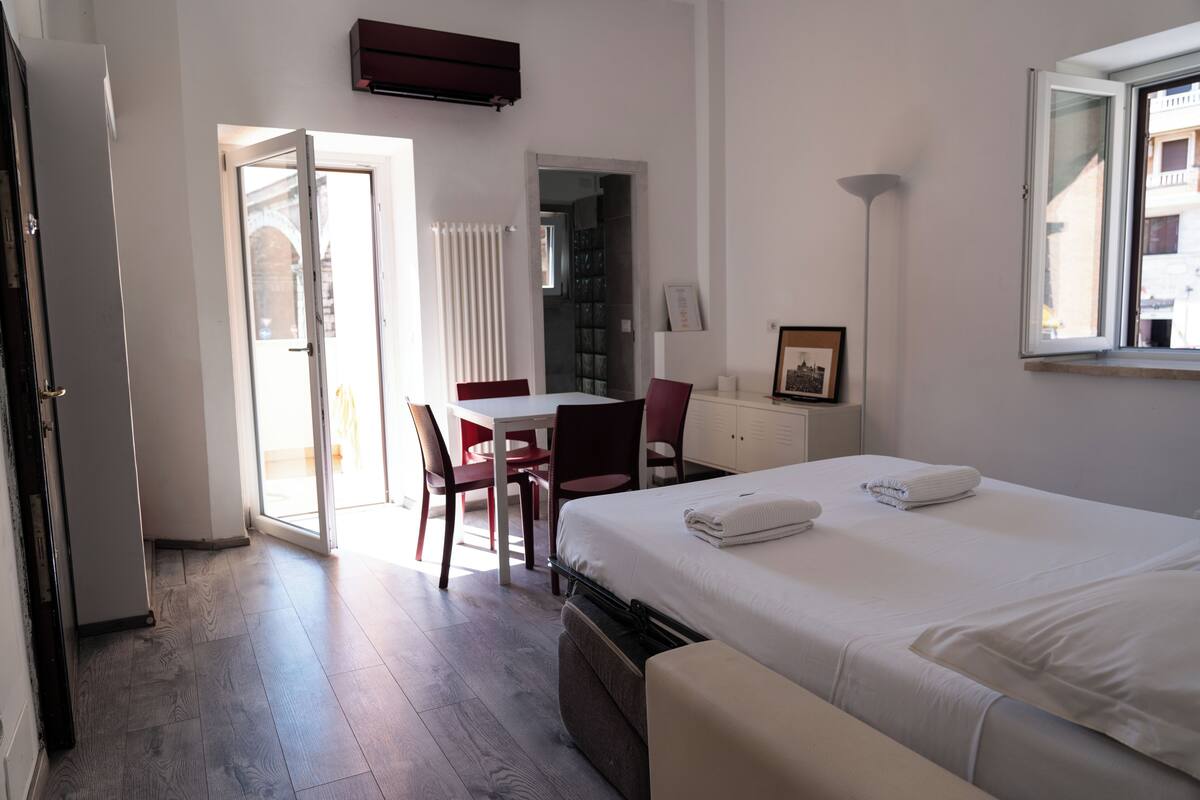 A hotel room with a bed, a desk, chairs, a radiator, a framed picture, and a window.
