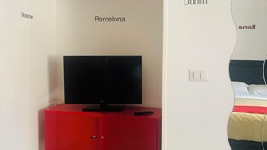 Habitació amb un suport de televisió vermell, un televisor de pantalla plana i un mirall que reflecteix un llit amb un llençol rayat vermell i blanc.