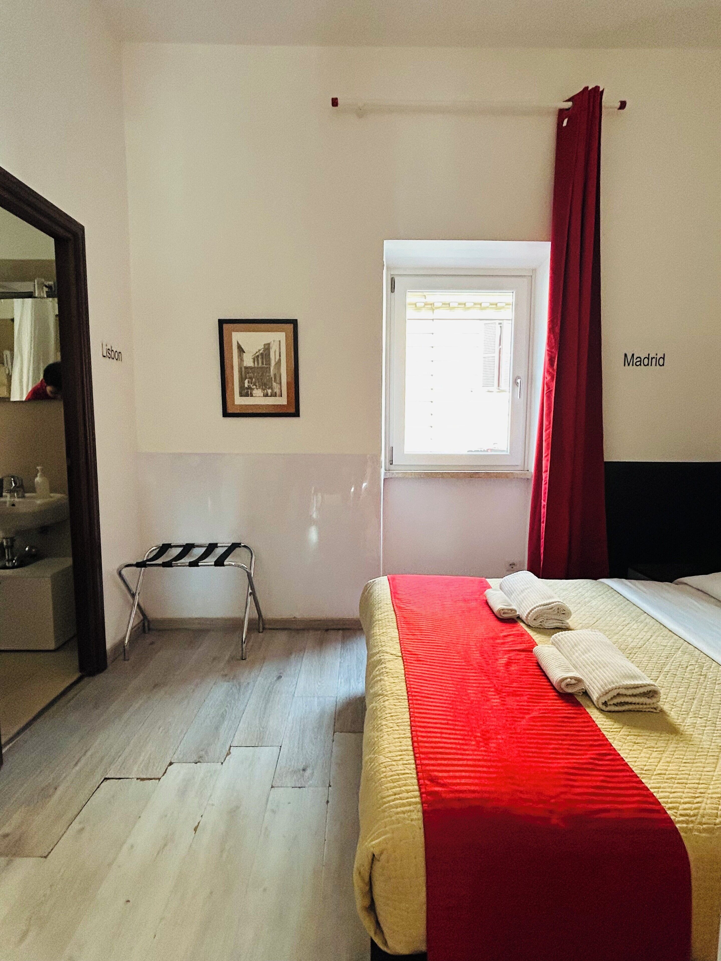 A bedroom with a bed, a window, a red curtain, a towel rack, and a framed picture on the wall.