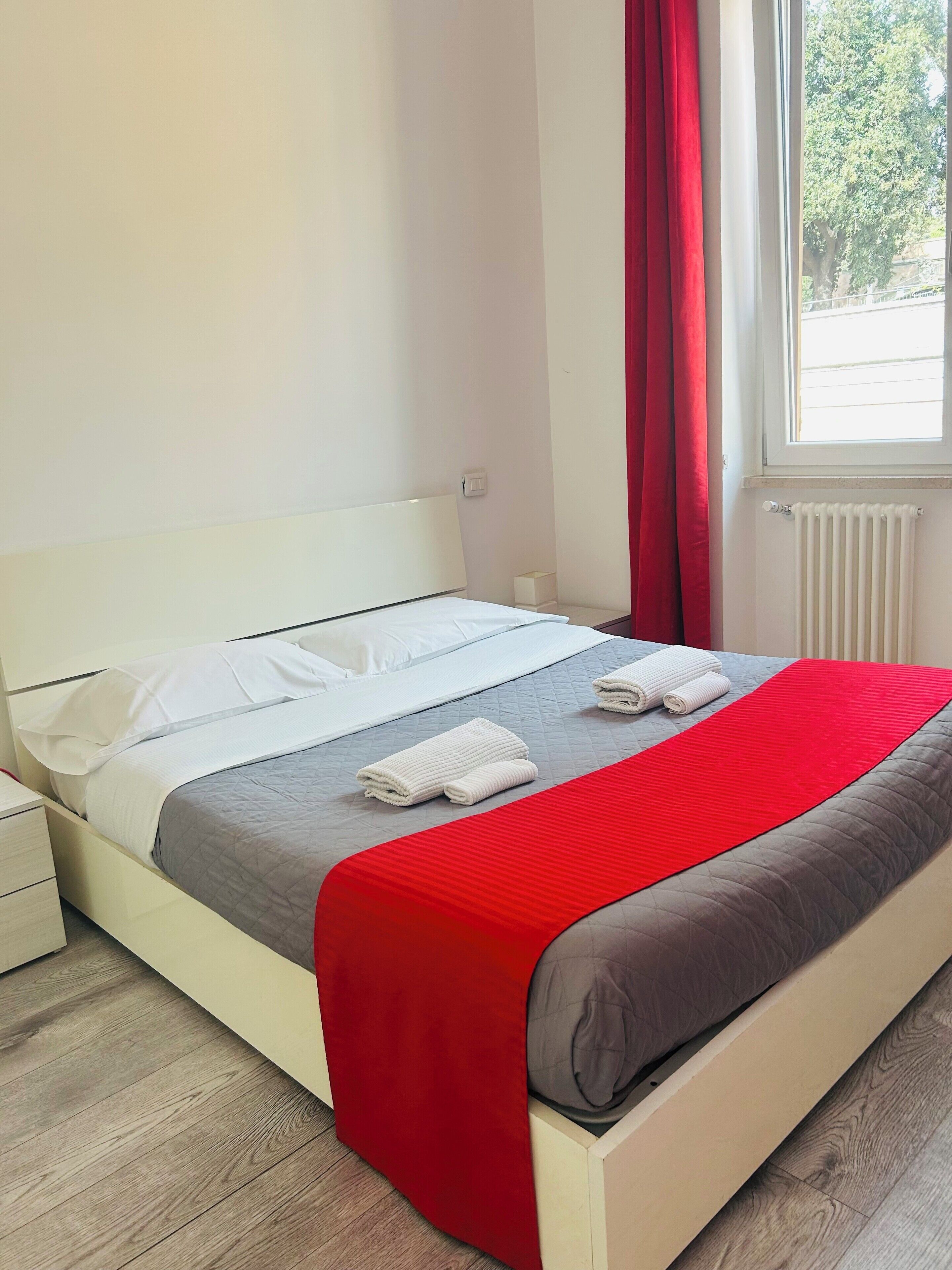 A neatly made bed with white and grey bedding, a red blanket, and two rolled towels.