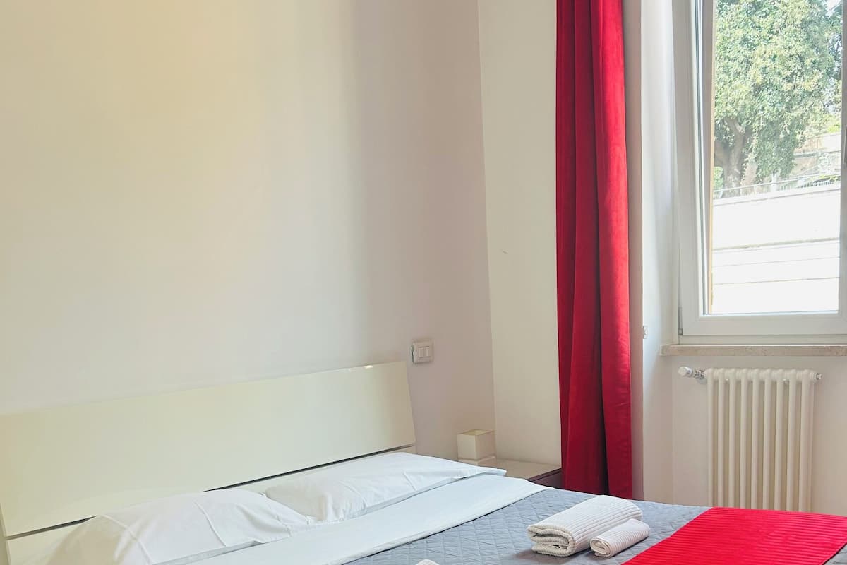 A neatly made bed with white and grey bedding, a red blanket, and two rolled towels.