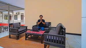 A man sitting on a chair with a laptop on a table in a room with a tiled floor and a beige wall. - OYO Life 92641 Guest House Palinggihan Ii (Sumber)