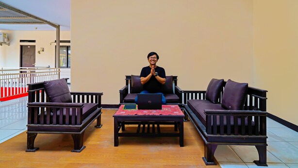 A person sitting on a dark-colored sofa in a room with a tiled floor and a plain wall. - OYO Life 92641 Guest House Palinggihan Ii (Sumber)