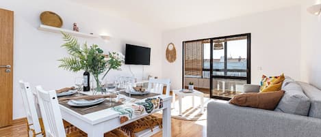 A modern living room with a dining area, a grey sofa, and a large window offering a view.