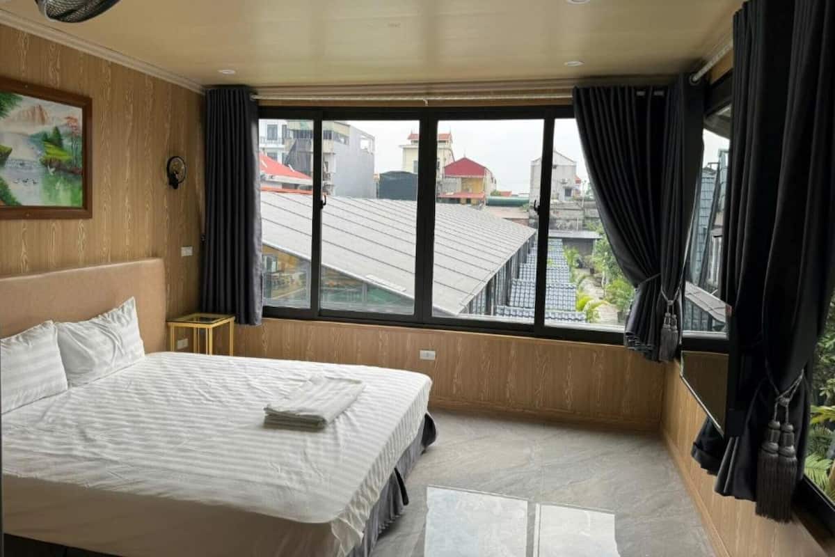 Hotellihuone, jossa on sänky, pieni pöytä, ikkuna verhoilla ja taulu seinällä.