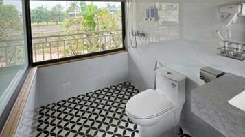 A modern bathroom with a toilet, sink, and shower. It features a geometric patterned floor and a view of greenery outside.