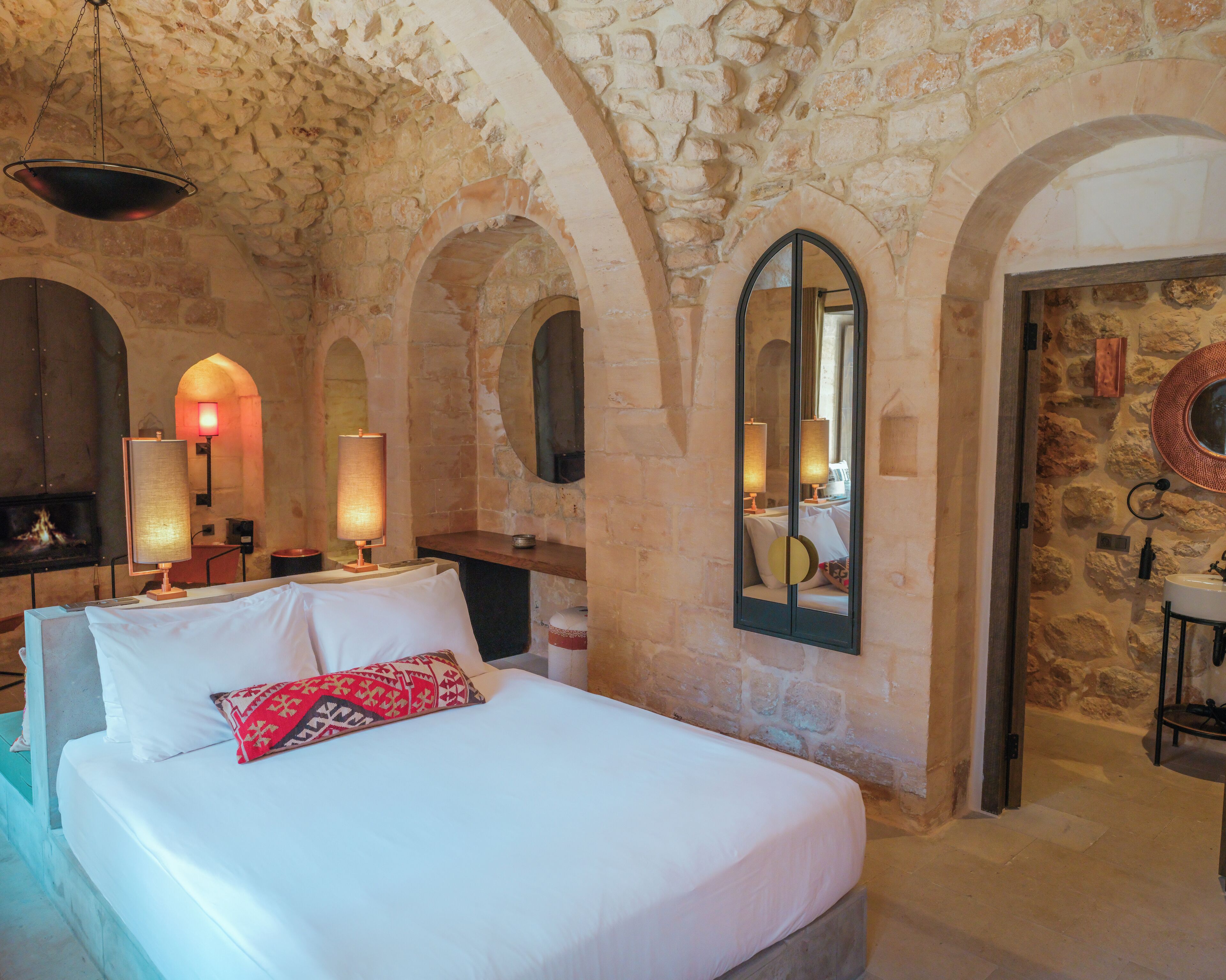 A room with a stone wall, a bed with white bedding and a red pillow, a mirror, and a fireplace.