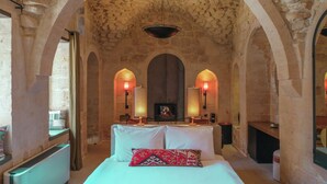 Una habitación con techo abovedado de piedra, cama con ropa de cama blanca y almohada estampada de color rojo, dos lámparas de pared y una chimenea.