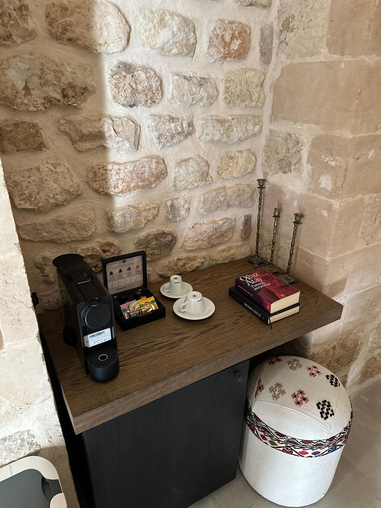 A small, stone-walled room with a wooden table holding a coffee maker, two cups, a box of chocolates, a book, and a decorative candle holder.