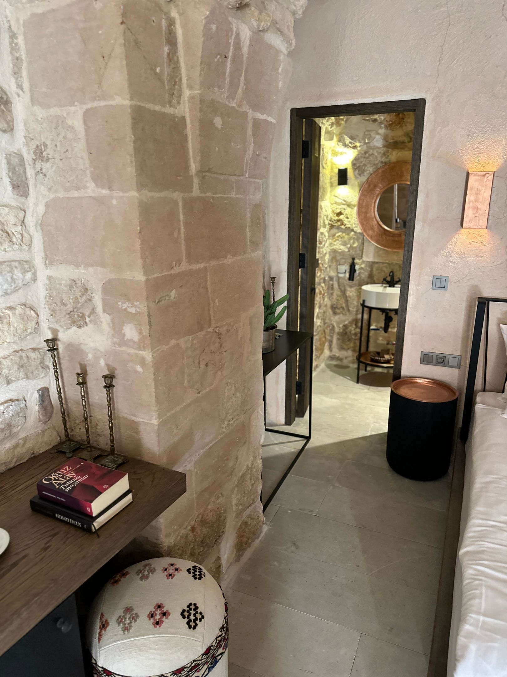 Un pasillo estrecho con paredes de piedra, un banco de madera con libros, un reposapiés estampado y un espejo con marco circular.