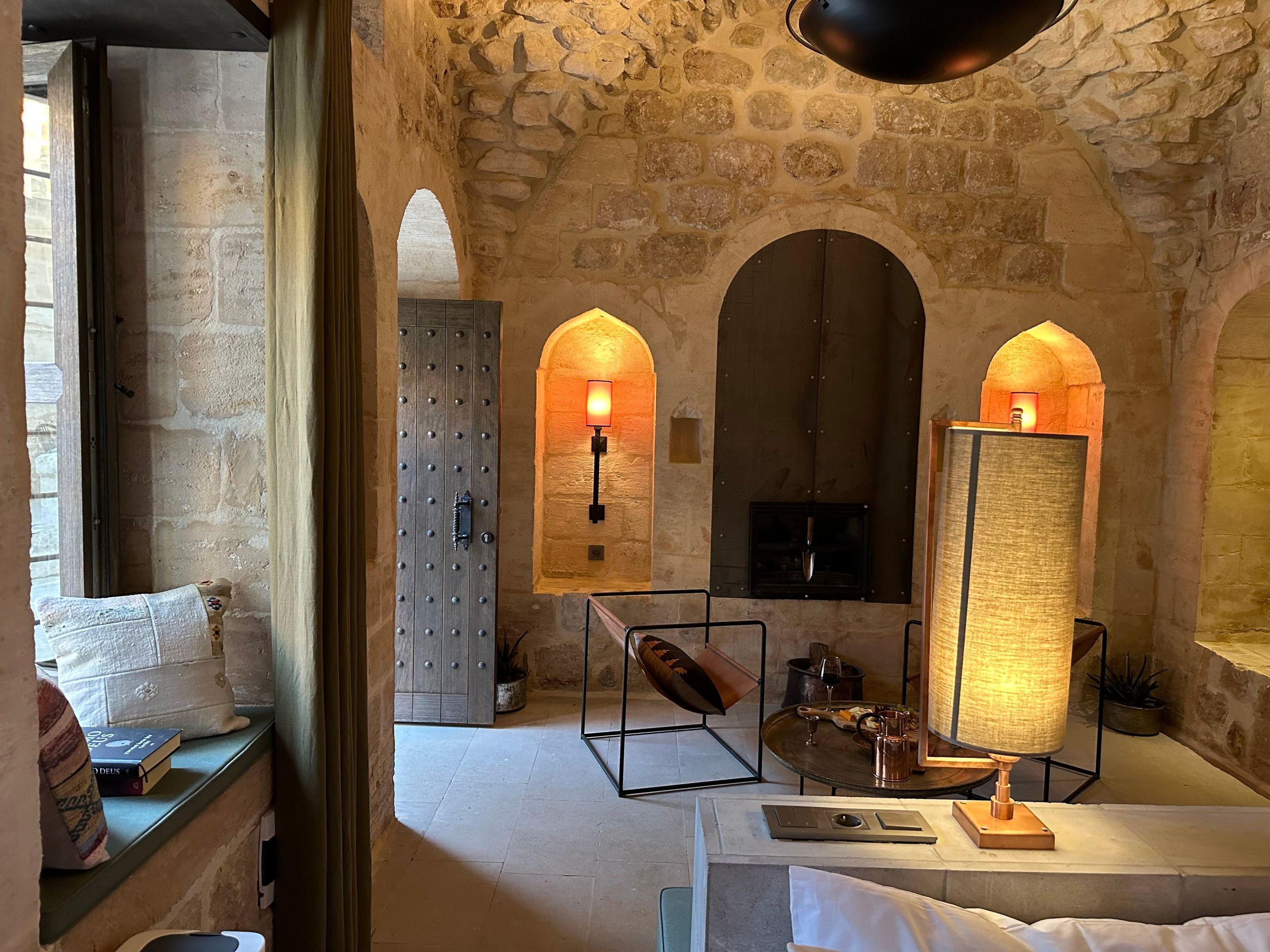 A stone-walled room with arched doorways, a fireplace, and a table with a lamp.