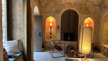 A stone-walled room with arched doorways, a fireplace, and a table with a lamp.