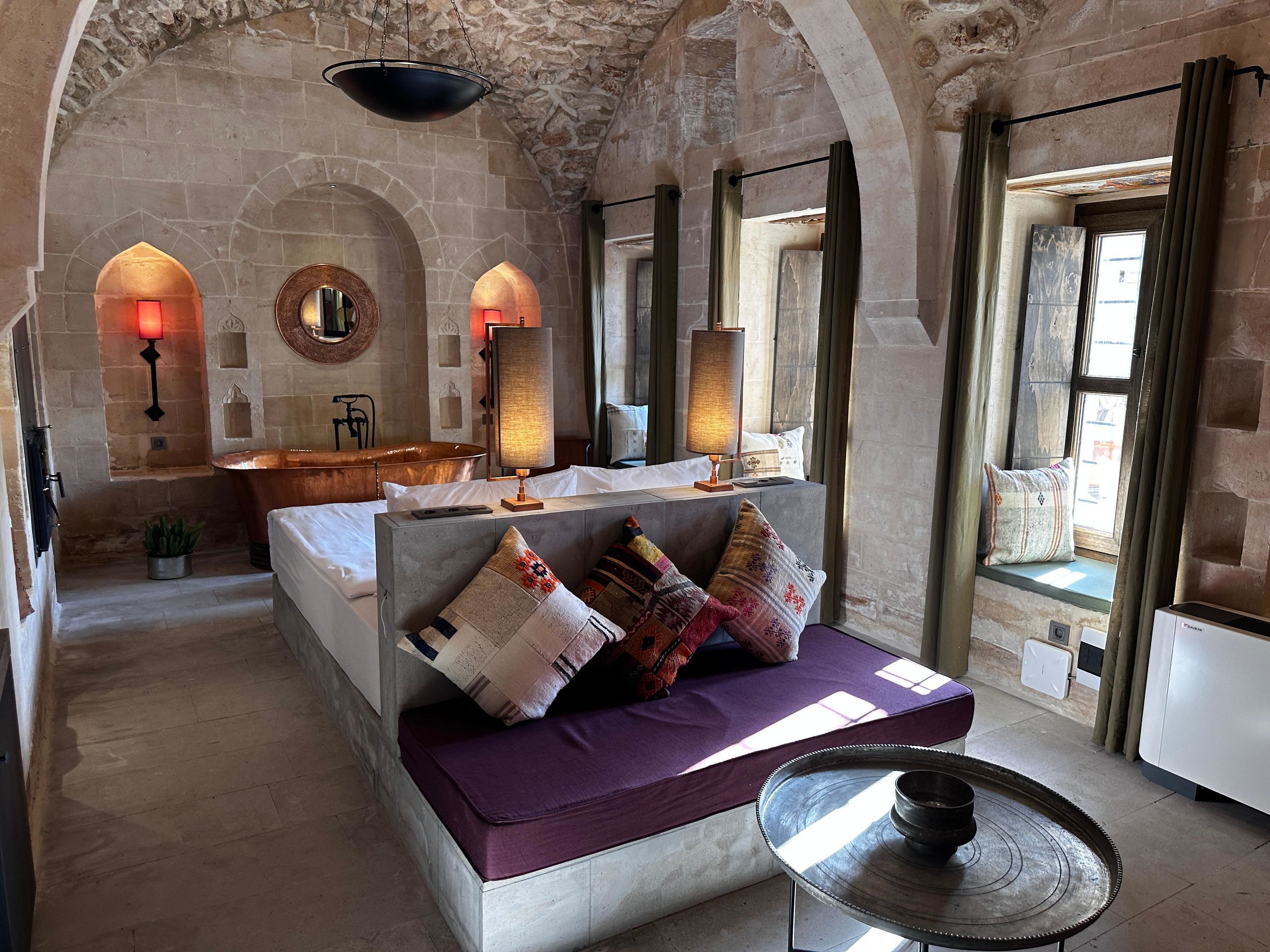 A cozy room with a stone wall, a large window, a bathtub, and a sofa with cushions.