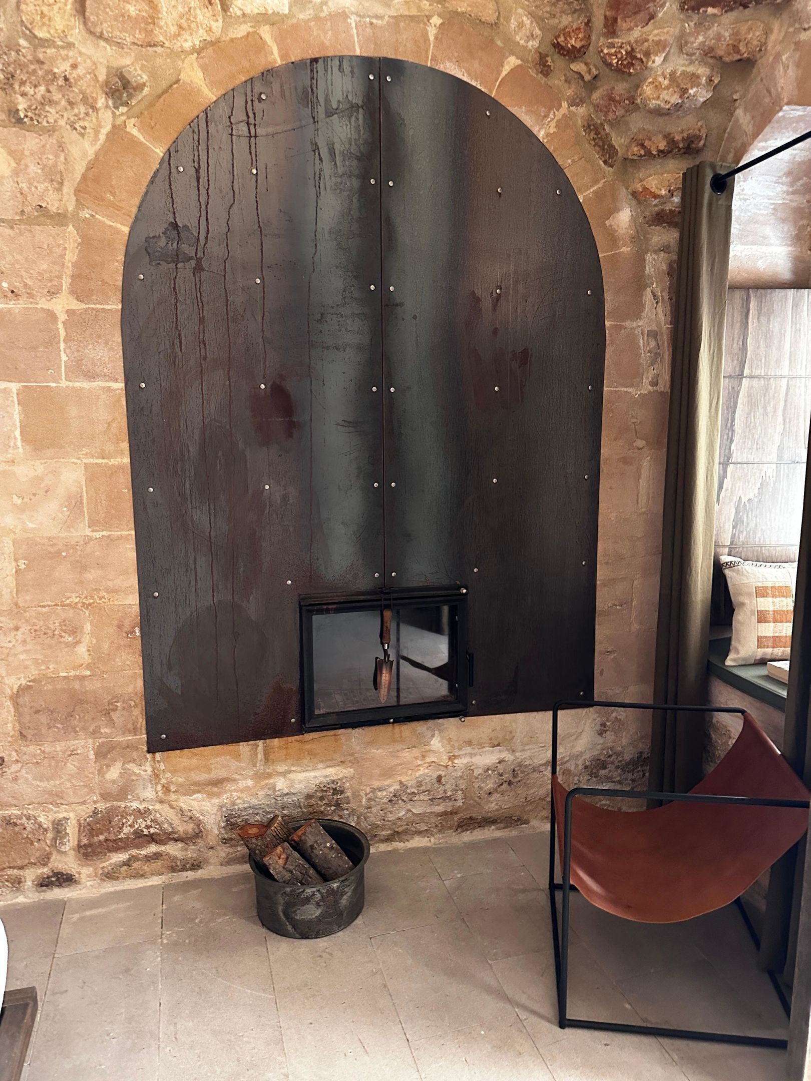 A rustic stone wall with a large, dark metal archway. Below the archway, there is a small black metal box with a handle. To the right, there is a red chair with a metal frame.