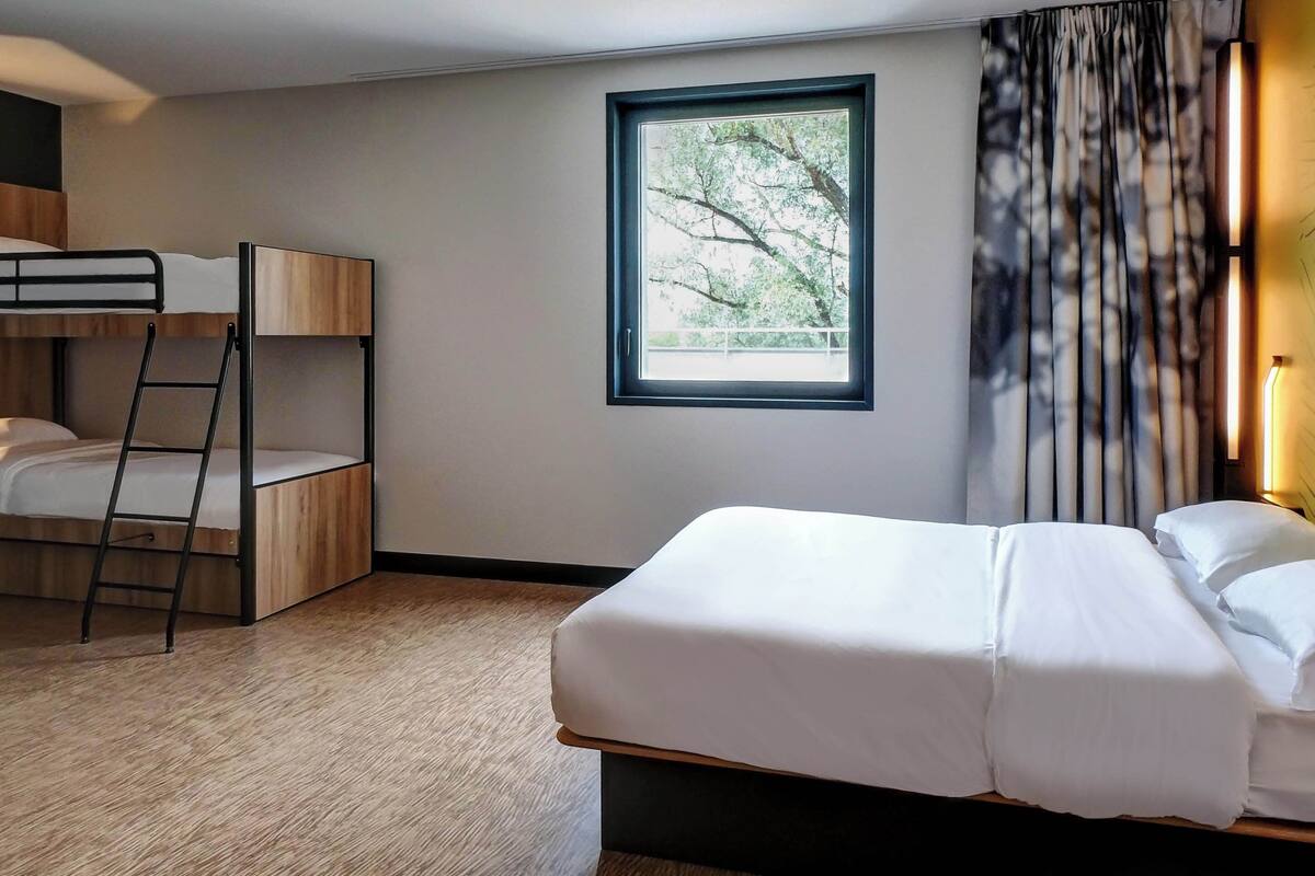 Ein Hotelzimmer mit einem Etagenbett, einem Einzelbett, einem Fenster mit Blick auf Bäume und einem Vorhang.