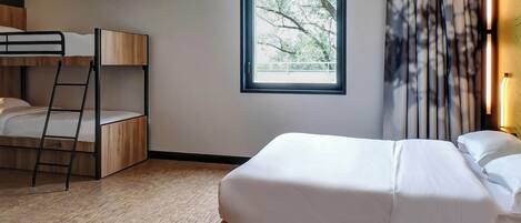 A hotel room with a bunk bed, a single bed, a window with a view of trees, and a curtain.