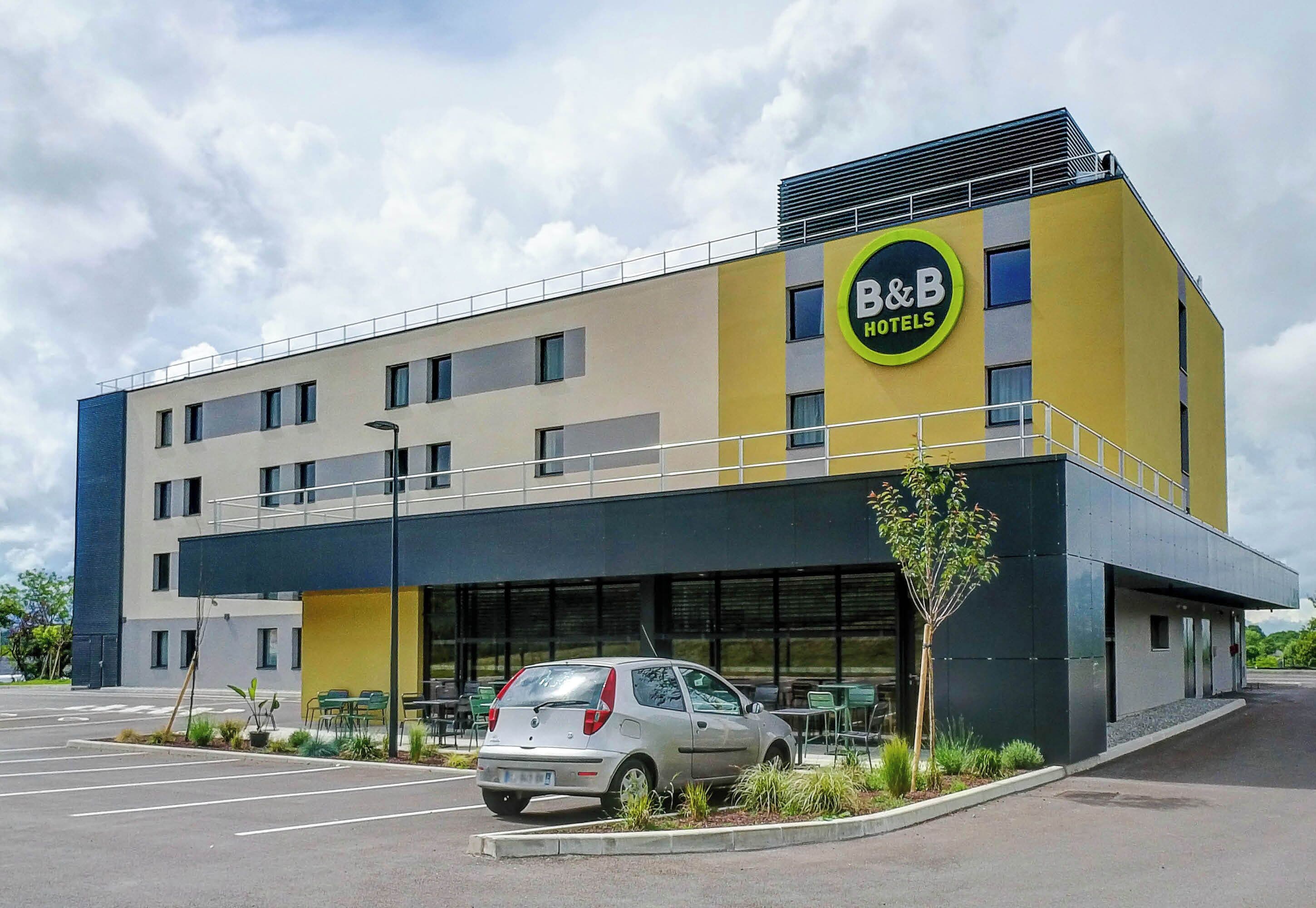 Un hotel B&B con diseño moderno, que presenta fachada amarilla y negra, un estacionamiento y un coche pequeño estacionado al frente.