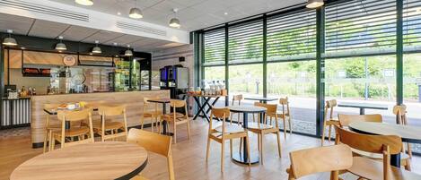 A modern cafe interior with wooden tables and chairs, a counter with a menu, and large windows offering a view of greenery outside.