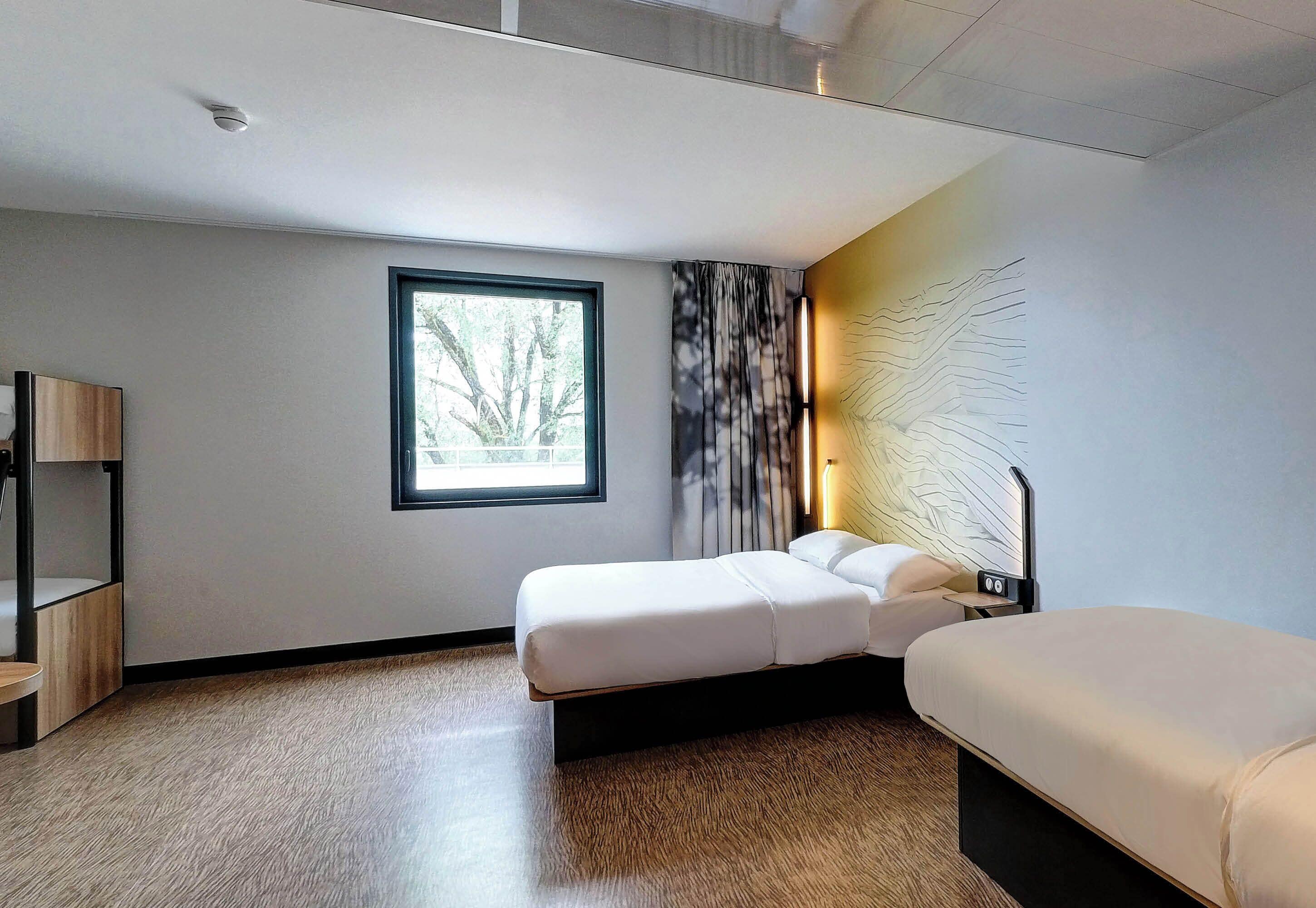 Una habitación de hotel moderna con un ventanal grande, una cama, una mesita de noche y una lámpara.