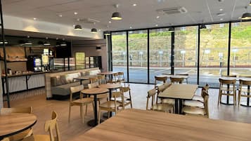Un café moderno con mesas y sillas de madera, una barra con un menú y amplios ventanales con vistas al exterior.