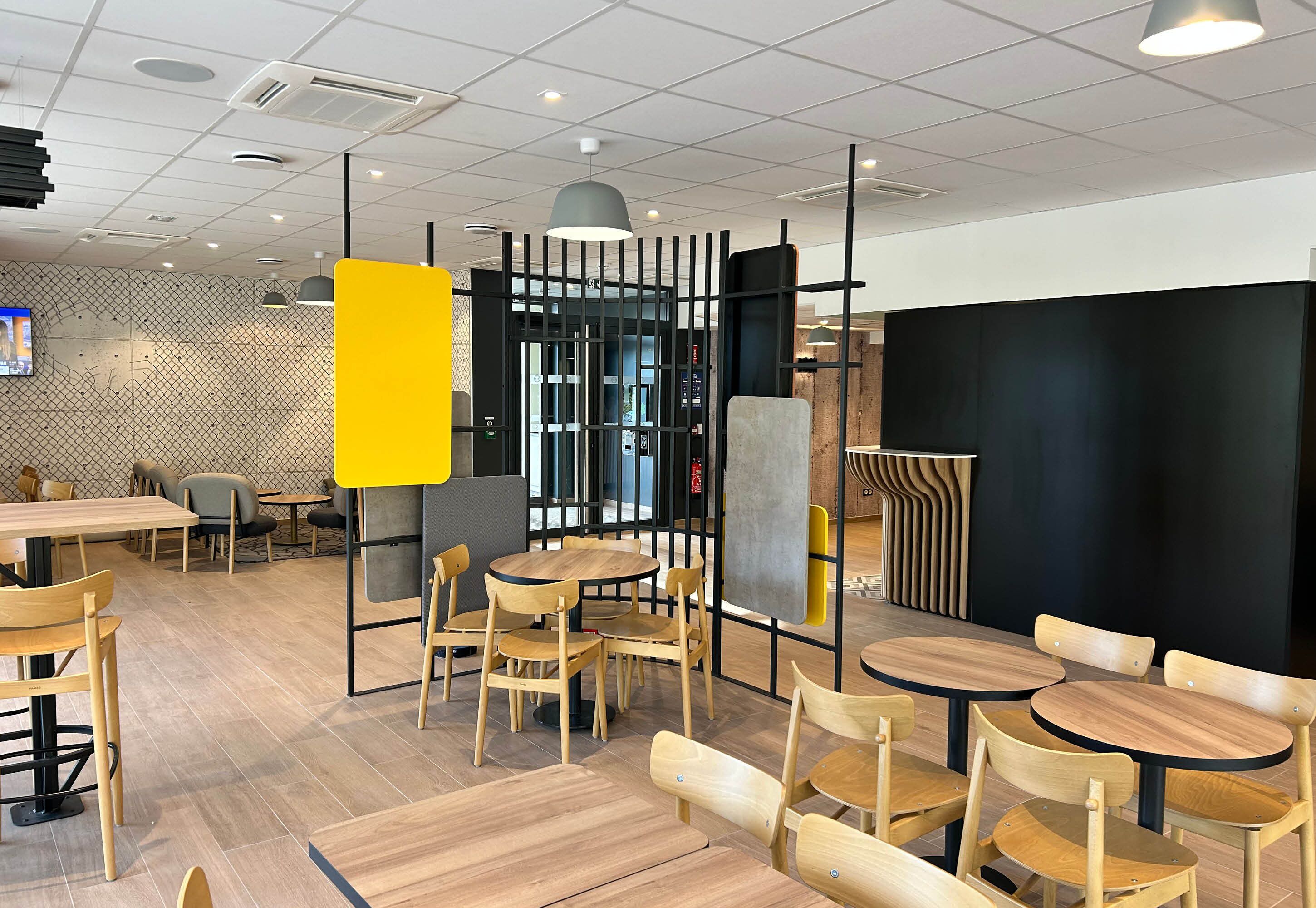 Un interior de restaurante moderno con mesas y sillas de madera, un separador amarillo y una pared de acento negra.