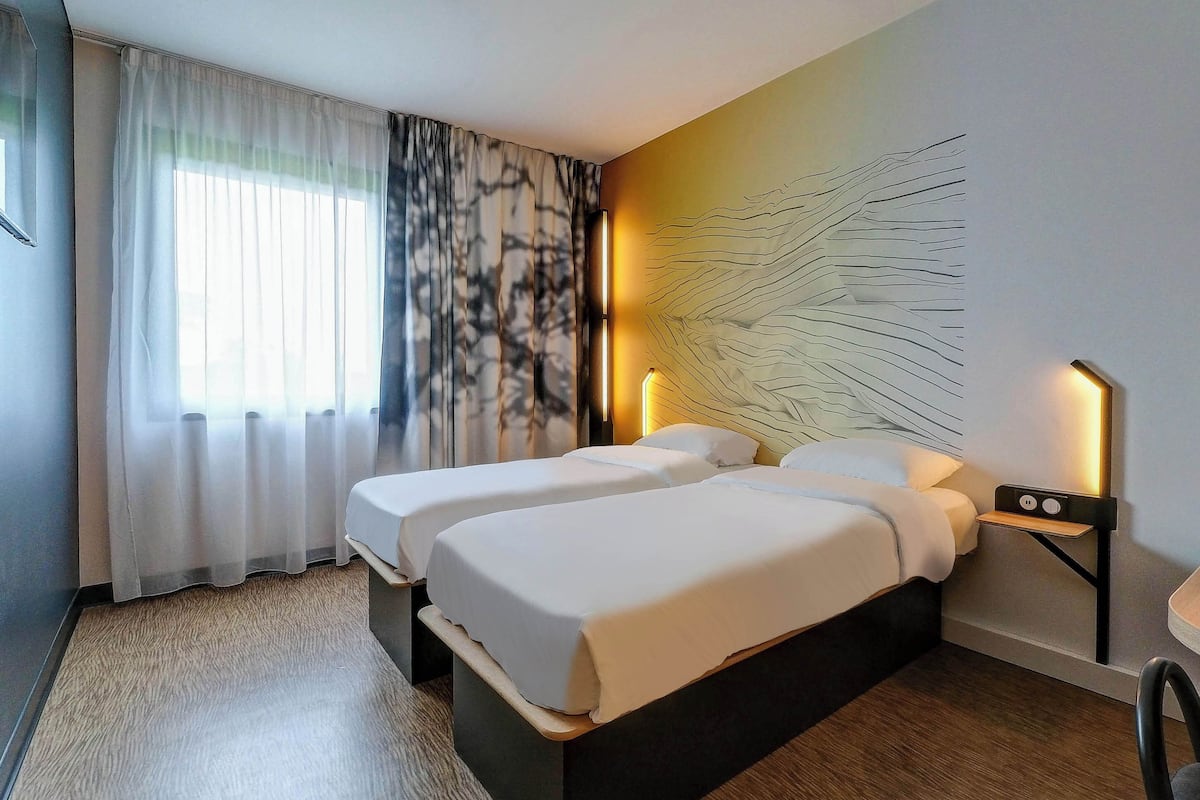 Ein Hotelzimmer mit zwei Betten, einem großen Fenster mit transparenten Vorhängen, einem Wandbild und Nachttischlampen.