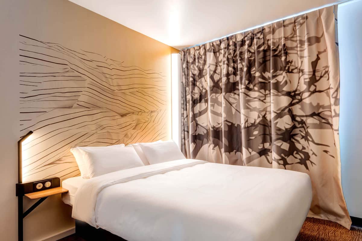 Ein modernes Hotelzimmer mit einem großen Bett, einer Kopfteil mit einem besonderen Lichtdesign, einem Nachttisch mit einer Lampe und einem Vorhang mit Baummuster.