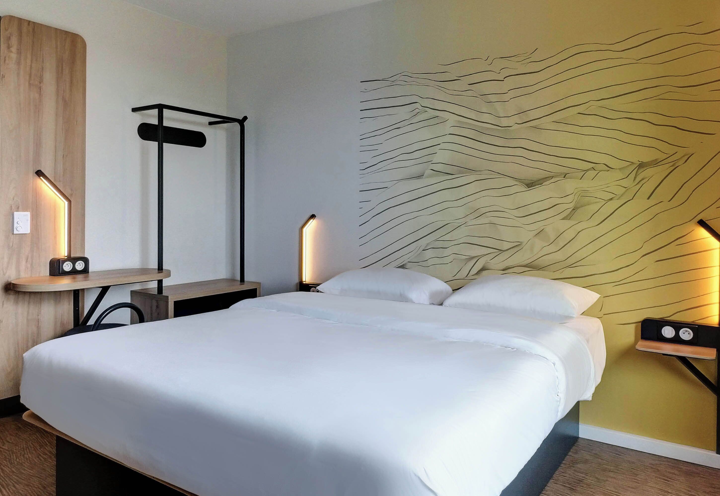 A modern hotel room with a large bed, a bedside table with a lamp, a wall-mounted light, and a unique textured wall design.