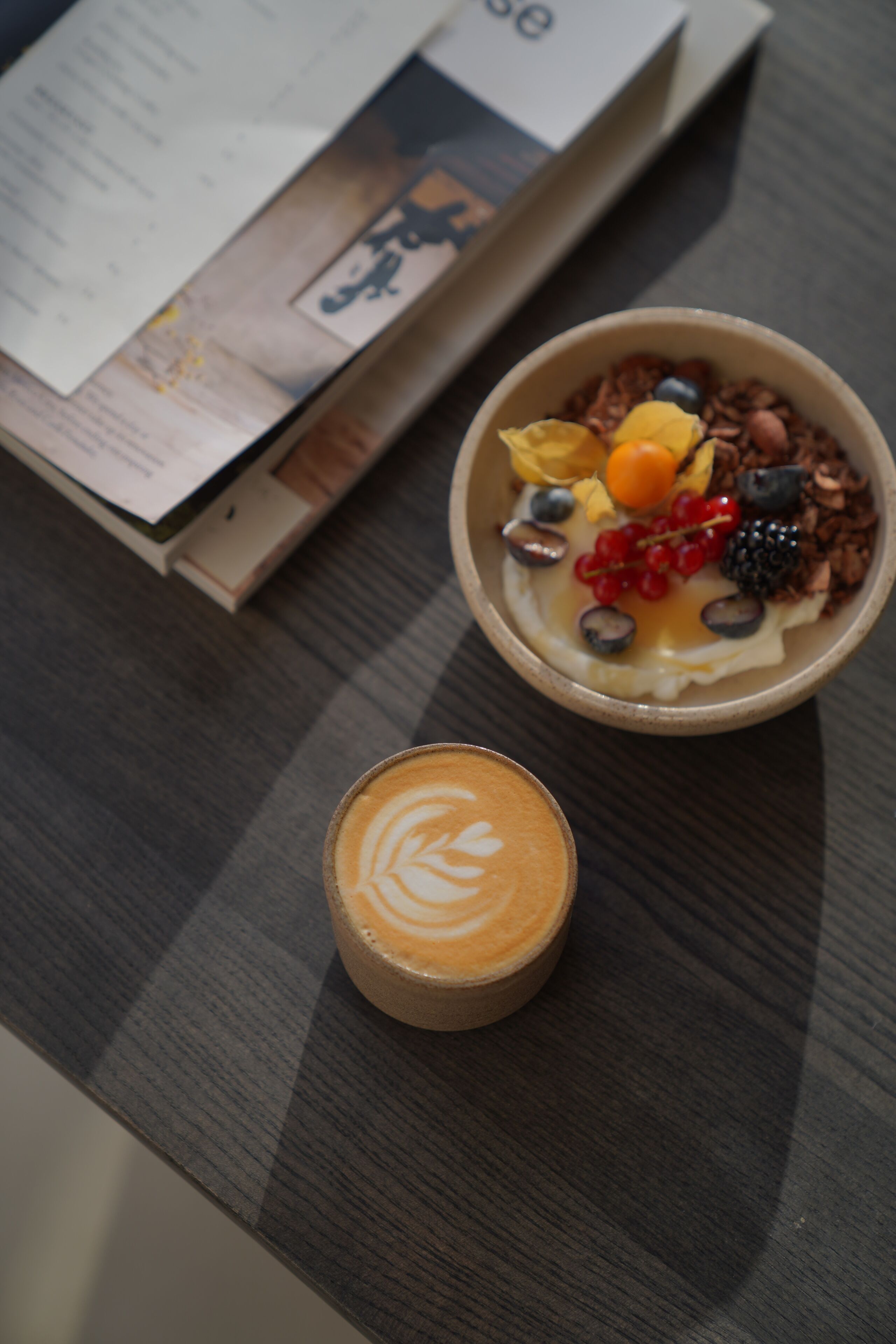 A bowl of yogurt topped with fruits and granola, a cup of coffee with latte art, and a menu on a table.