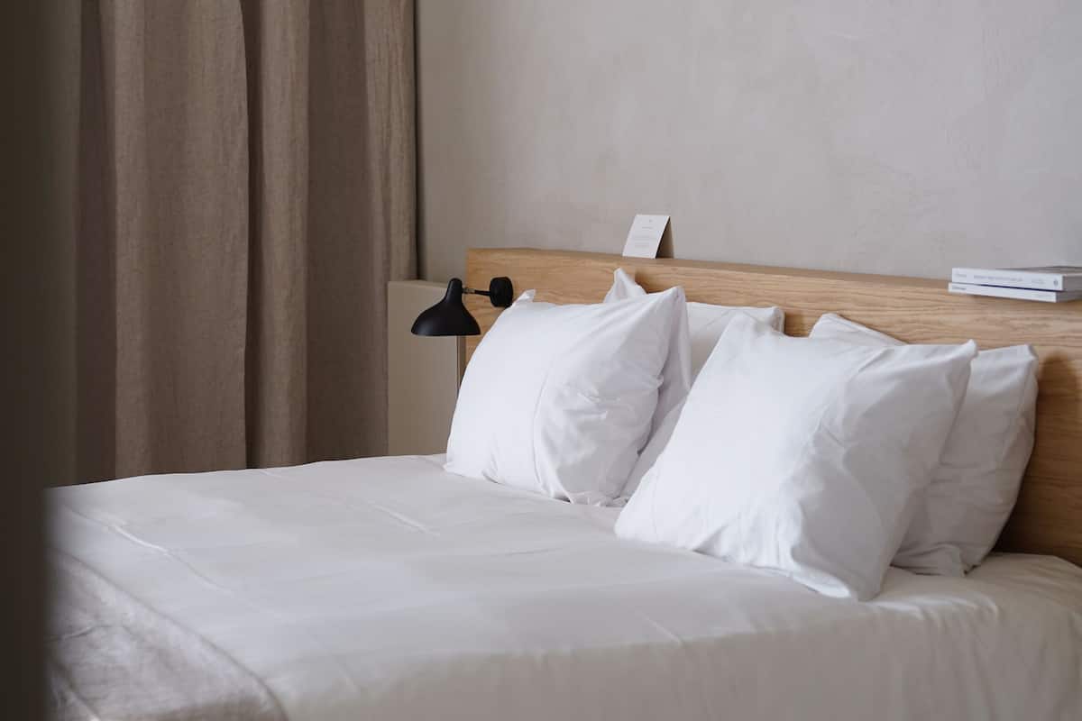 Una cama bien hecha con sábanas y almohadas blancas, un cabecero de madera y una lámpara negra a su lado.