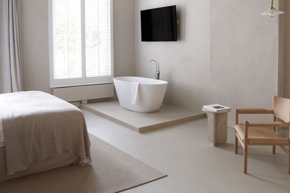 Un dormitorio moderno con una cama grande, una bañera independiente, un televisor montado en la pared y una silla de madera.