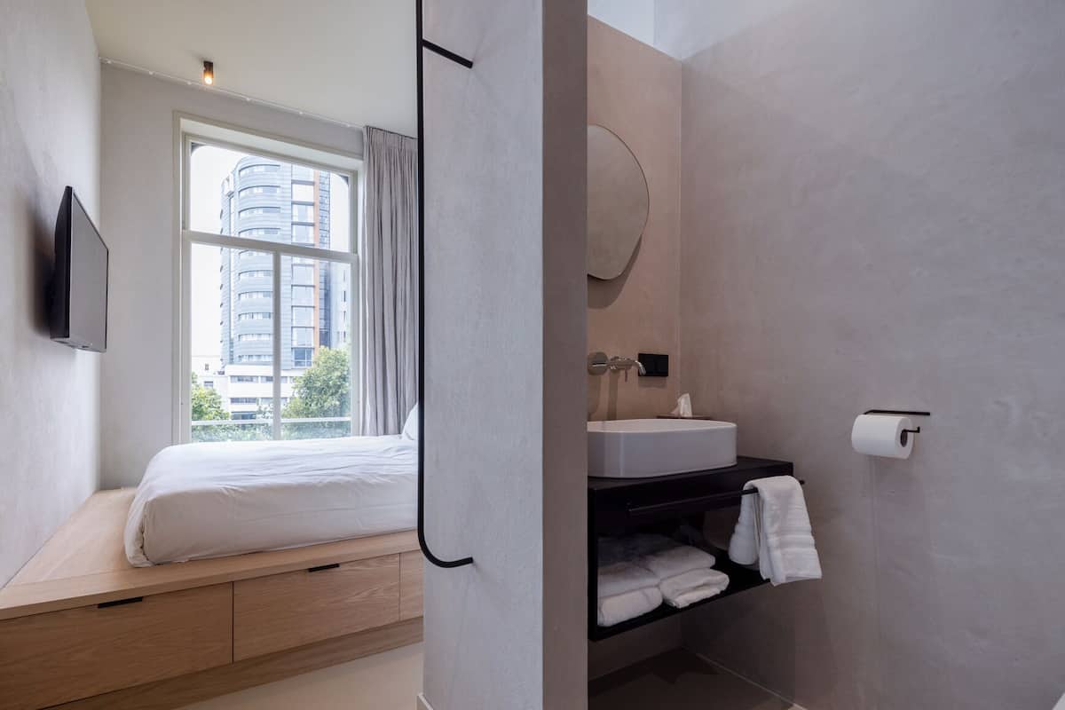 Una habitación de hotel moderna con cama, televisor, ventana con cortinas, baño con lavamanos, toallero y papel higiénico.