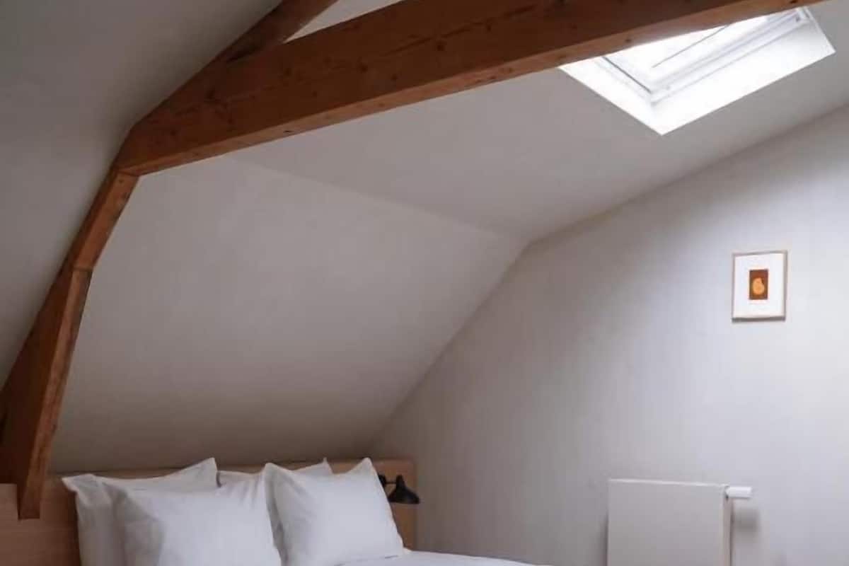 Un dormitorio con techo inclinado, tragaluz, cama con ropa de cama blanca y radiador.