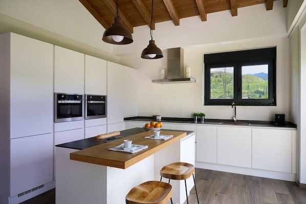 Eine moderne Küche mit weißen Schränken, einer Holz-Kochinsel, zwei Pendelleuchten und einem Fenster mit Blick auf Grünflächen.
