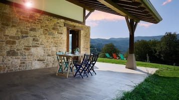 Un edificio de piedra con un porche cubierto, una mesa con sillas y vistas a montañas.