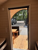 Uma porta aberta dá acesso a um pátio externo com uma mesa e um banco de madeira, rodeados por uma área de cascalho e árvores.