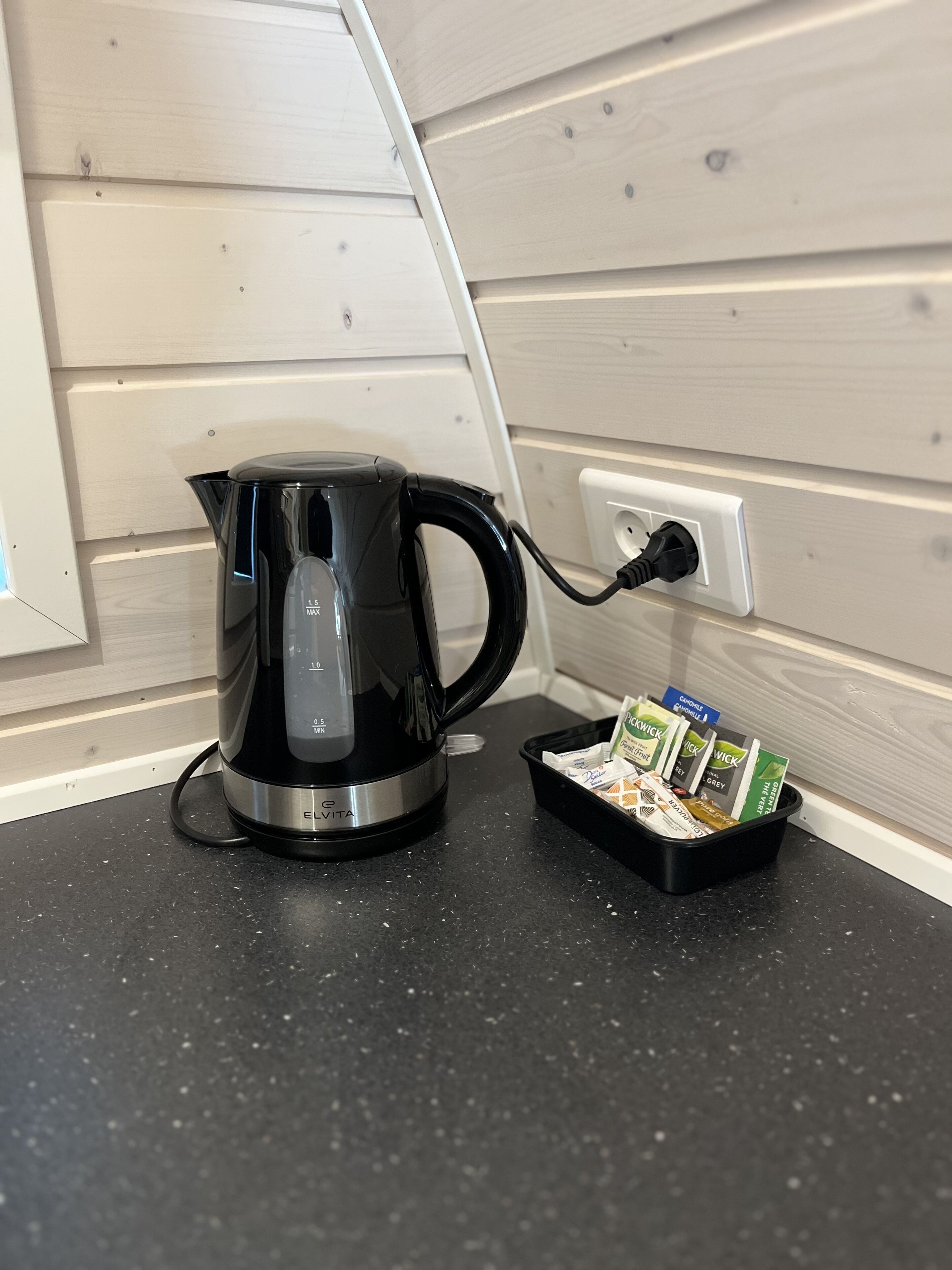 Ein schwarzer Wasserkocher steht auf einer schwarzen Arbeitsplatte neben einer Steckdose, mit einer Ablage für Kaffeekapseln.