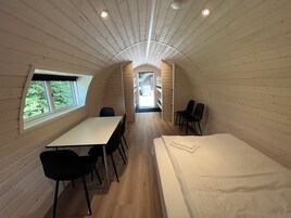 Quarto compacto estilo cabine, com uma mesa comprida e cadeiras, uma cama e uma janela com vista para vegetação.