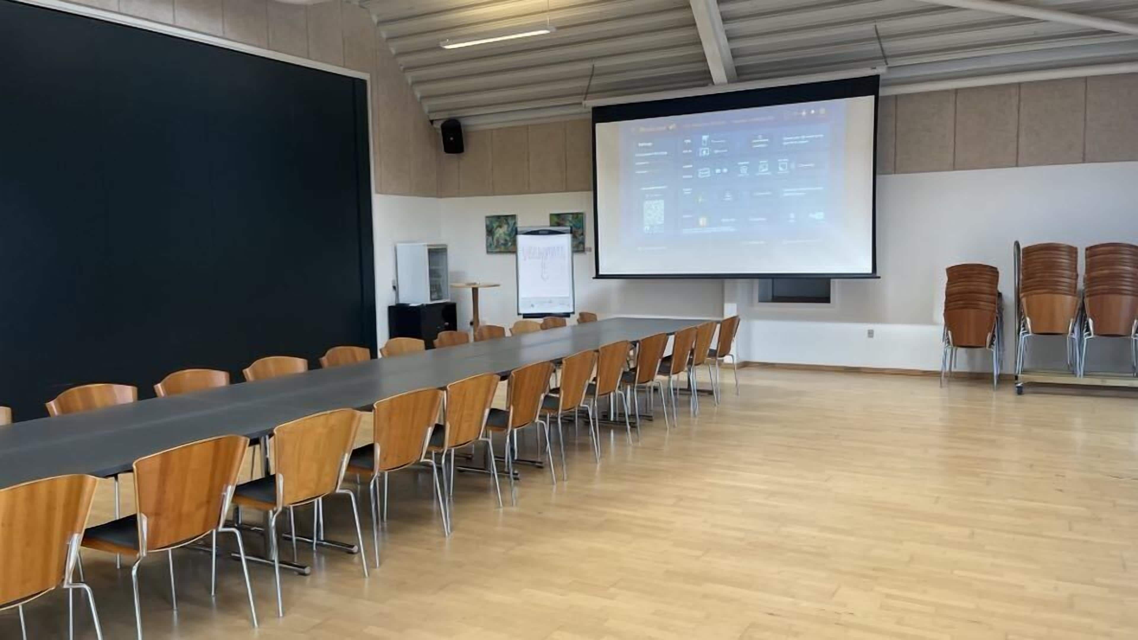 Ein Konferenzraum mit einem langen Tisch, Stühlen, einer Projektionsleinwand und einer Whiteboard-Tafel.