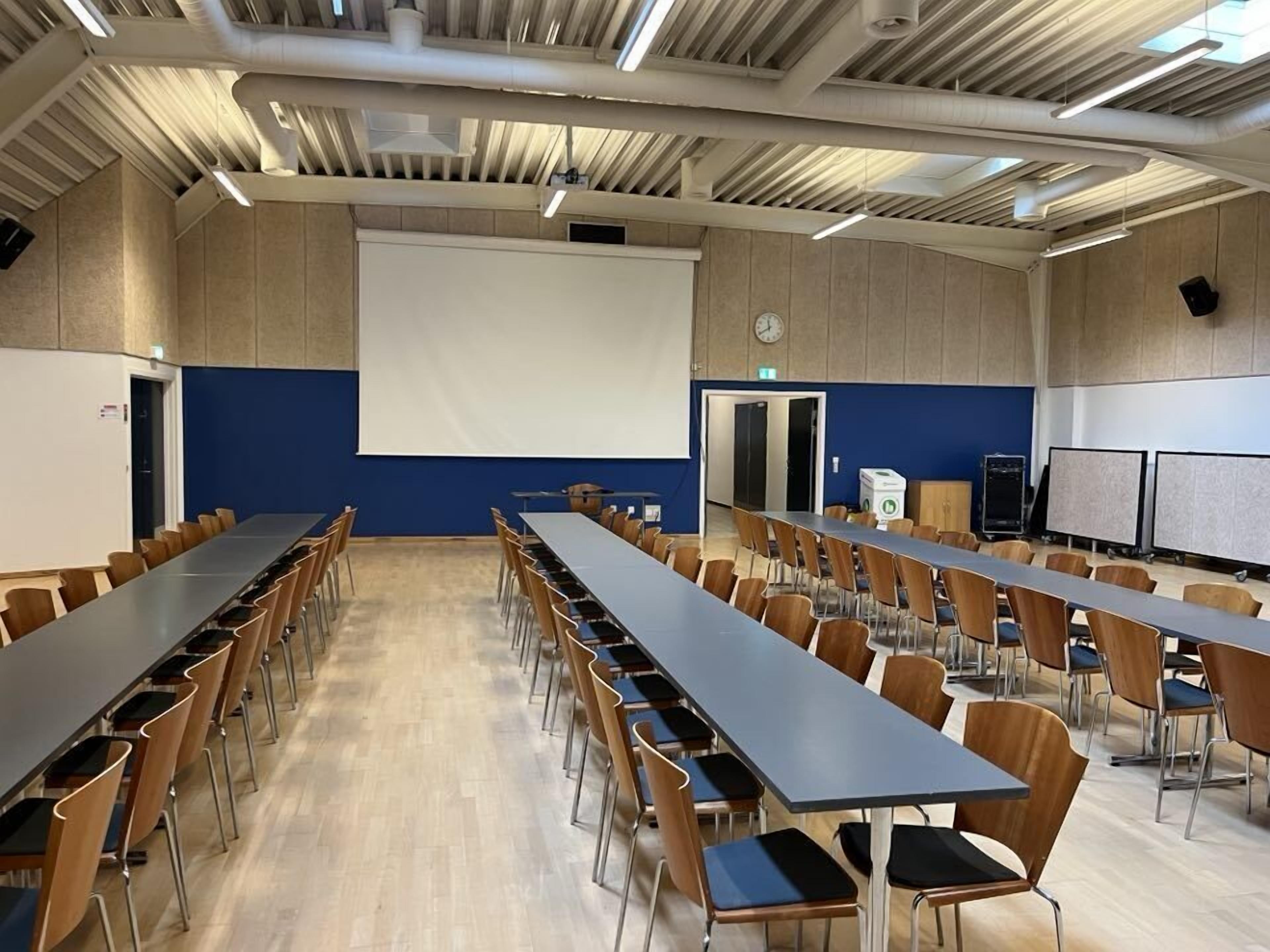 A conference room with long tables and chairs, a large screen, and a clock on the wall.
