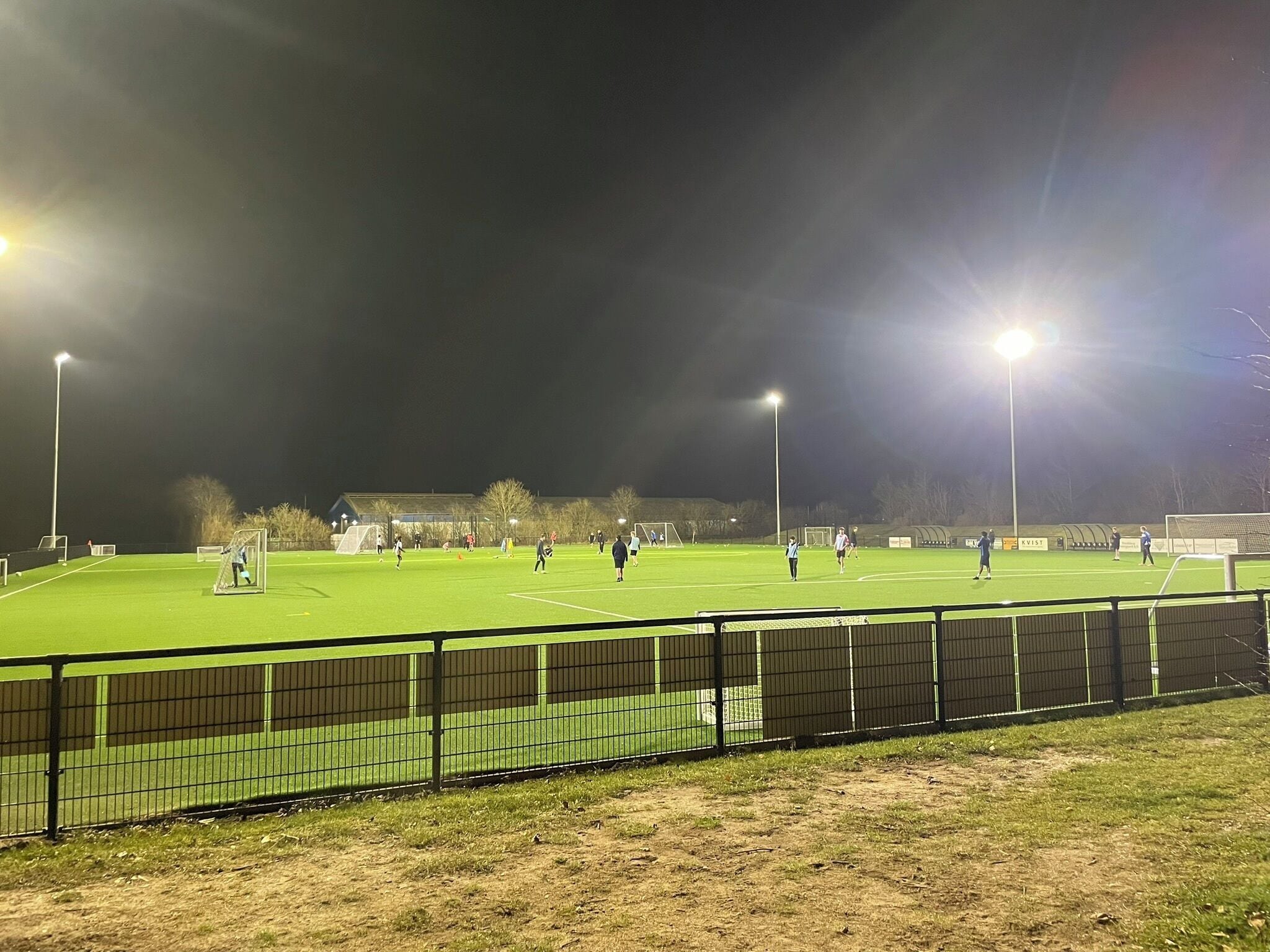 A well-lit sports field with goalposts, a fence, and a building in the background.