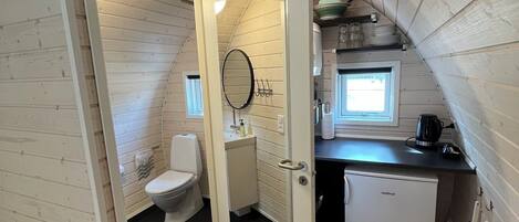 Une petite pièce de style cabane, construite en bois, comprenant des toilettes, un lavabo et un petit réfrigérateur.