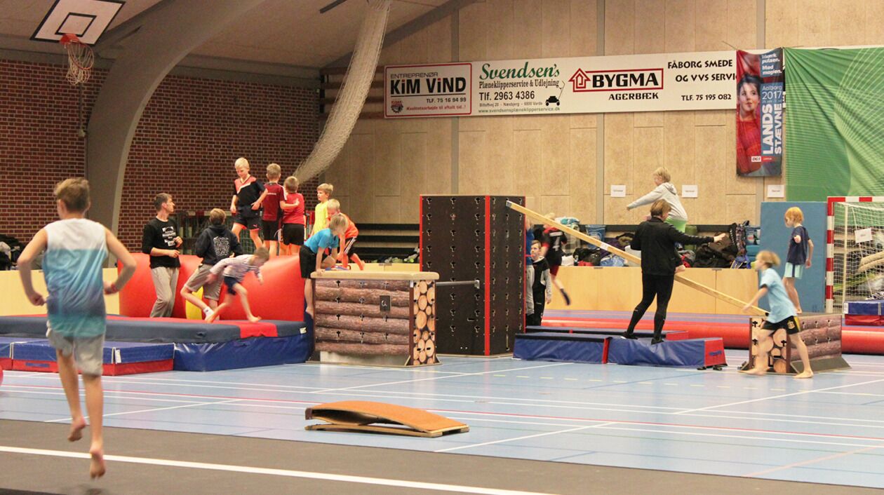 A gymnasium with a basketball hoop, gymnastics equipment, and a group of people engaged in activities.