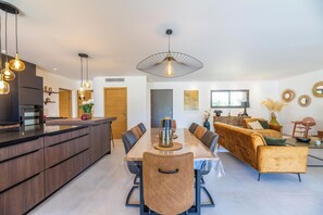 Une cuisine moderne dotée d’un îlot central, d’un coin repas avec une longue table et des chaises, et d’un espace de vie comprenant un canapé et des fauteuils.