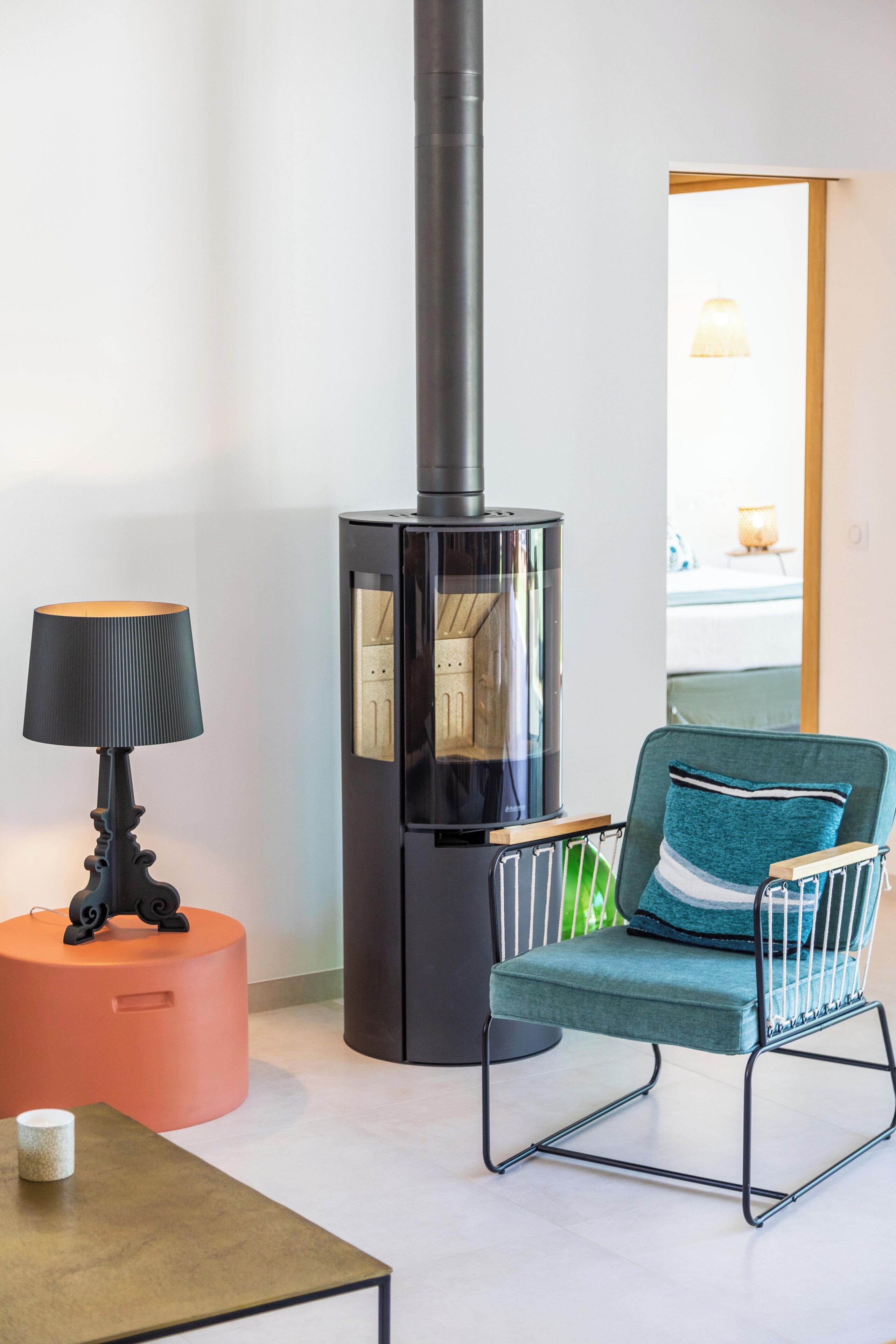 A modern living room with a black wood-burning stove, a teal chair with a striped cushion, a round orange ottoman, and a table with a lamp.
