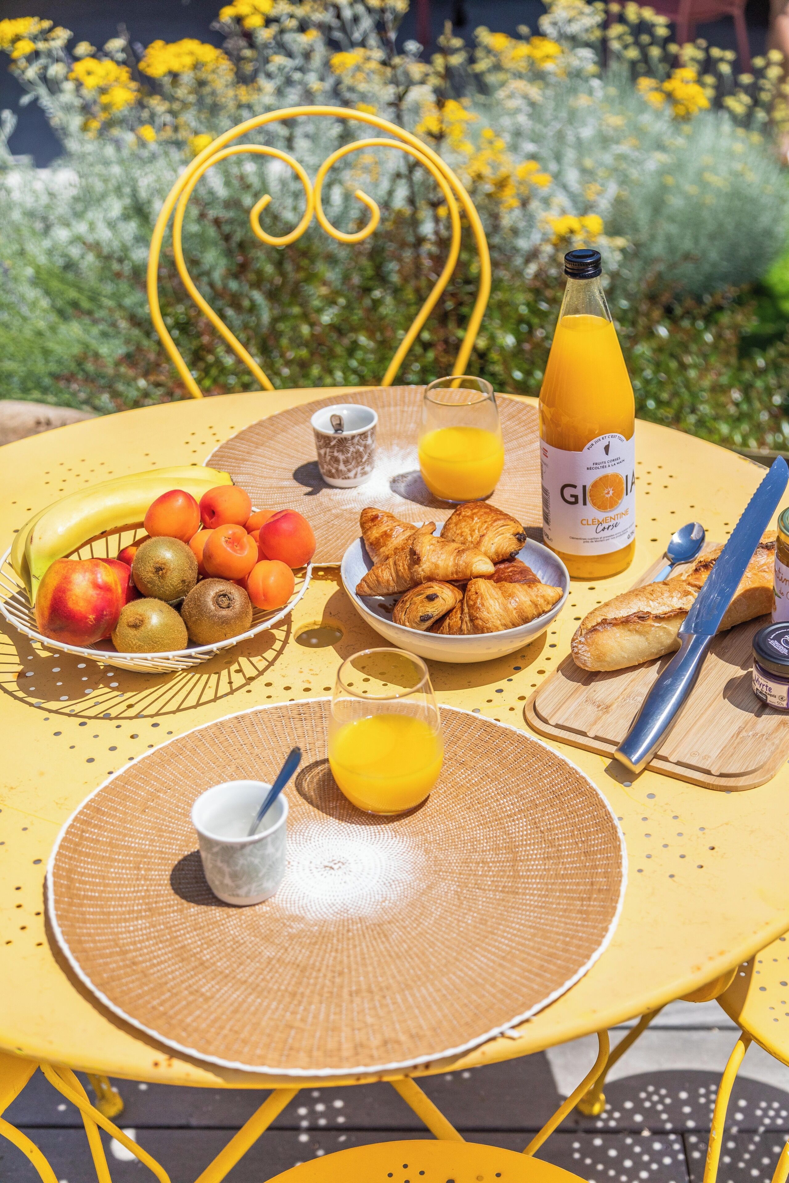 Ein Tisch mit Frühstücksangeboten, darunter Obst, Croissants und Saft, draußen mit einem gelben Stuhl und einem herzförmigen Metallrahmen im Hintergrund.