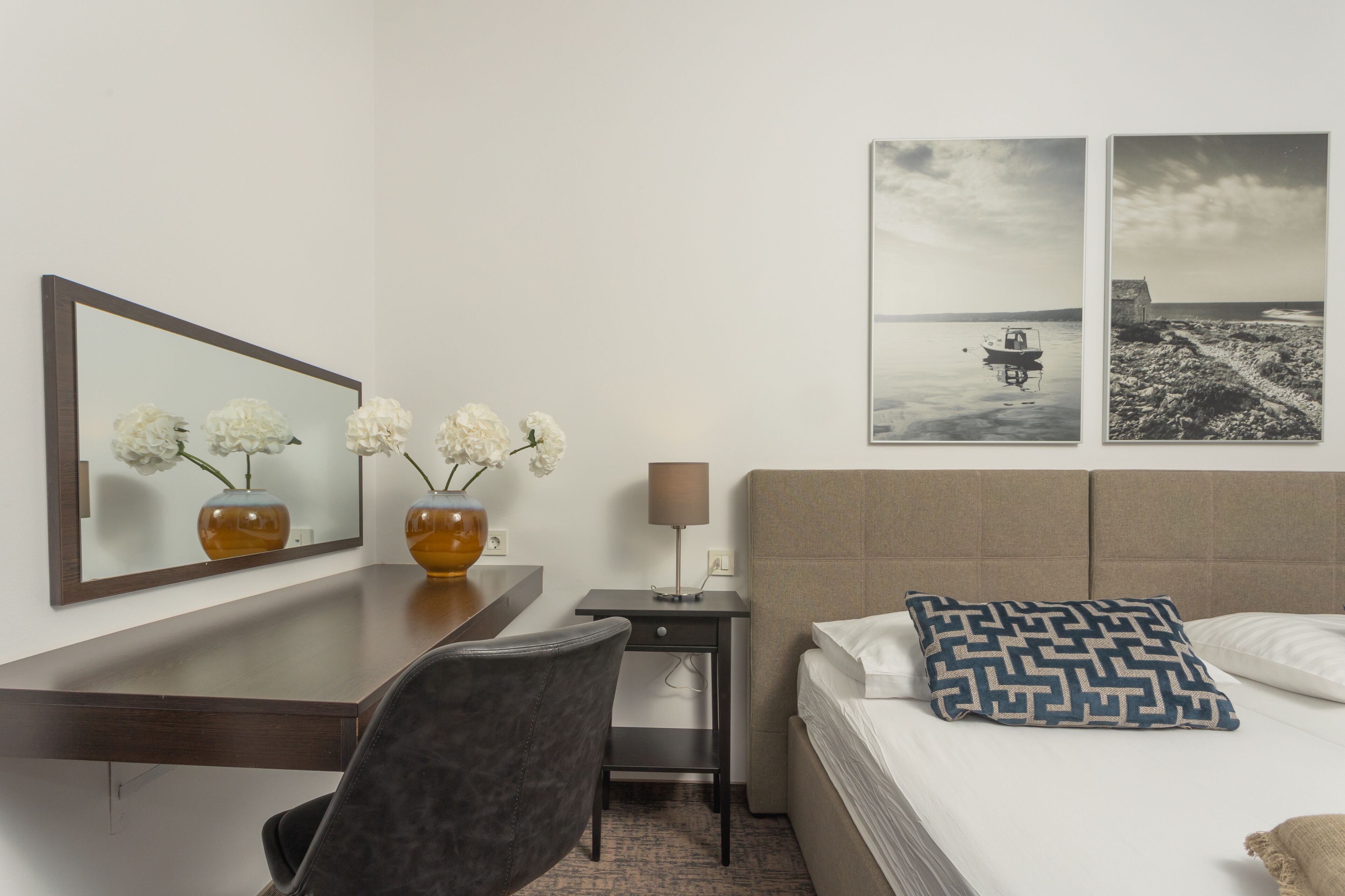 A hotel room with a bed, a desk, a chair, a lamp, and two framed pictures on the wall.